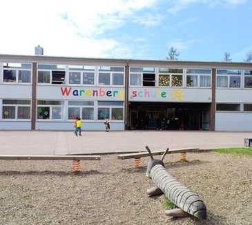 Die Warenbergschule ist ein zweigeschossiges Gebäude mit bunten Fensterdekorationen. Vor dem Eingang spielen Kinder.